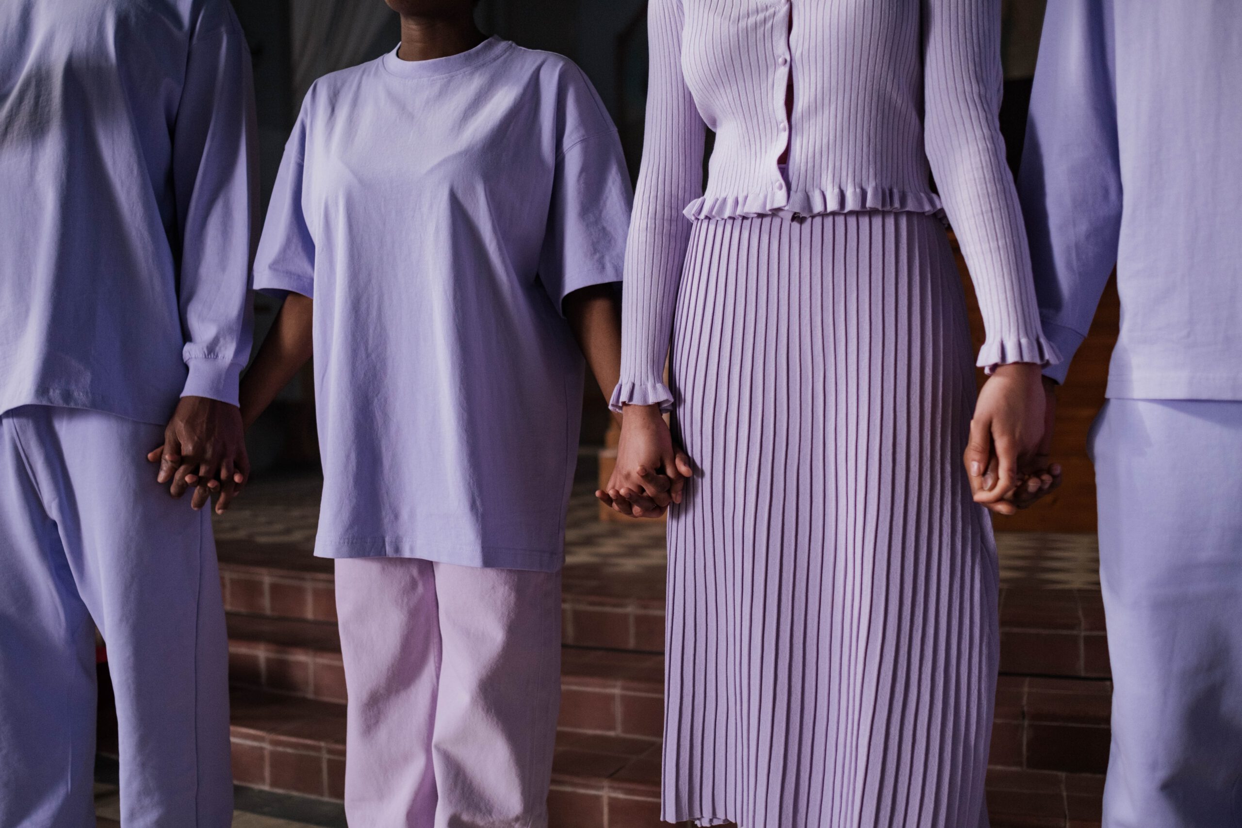 Ludzie ubrani na fioletowo trzymający się za ręce a wśród nich kobieta w plisowanej spódnicy