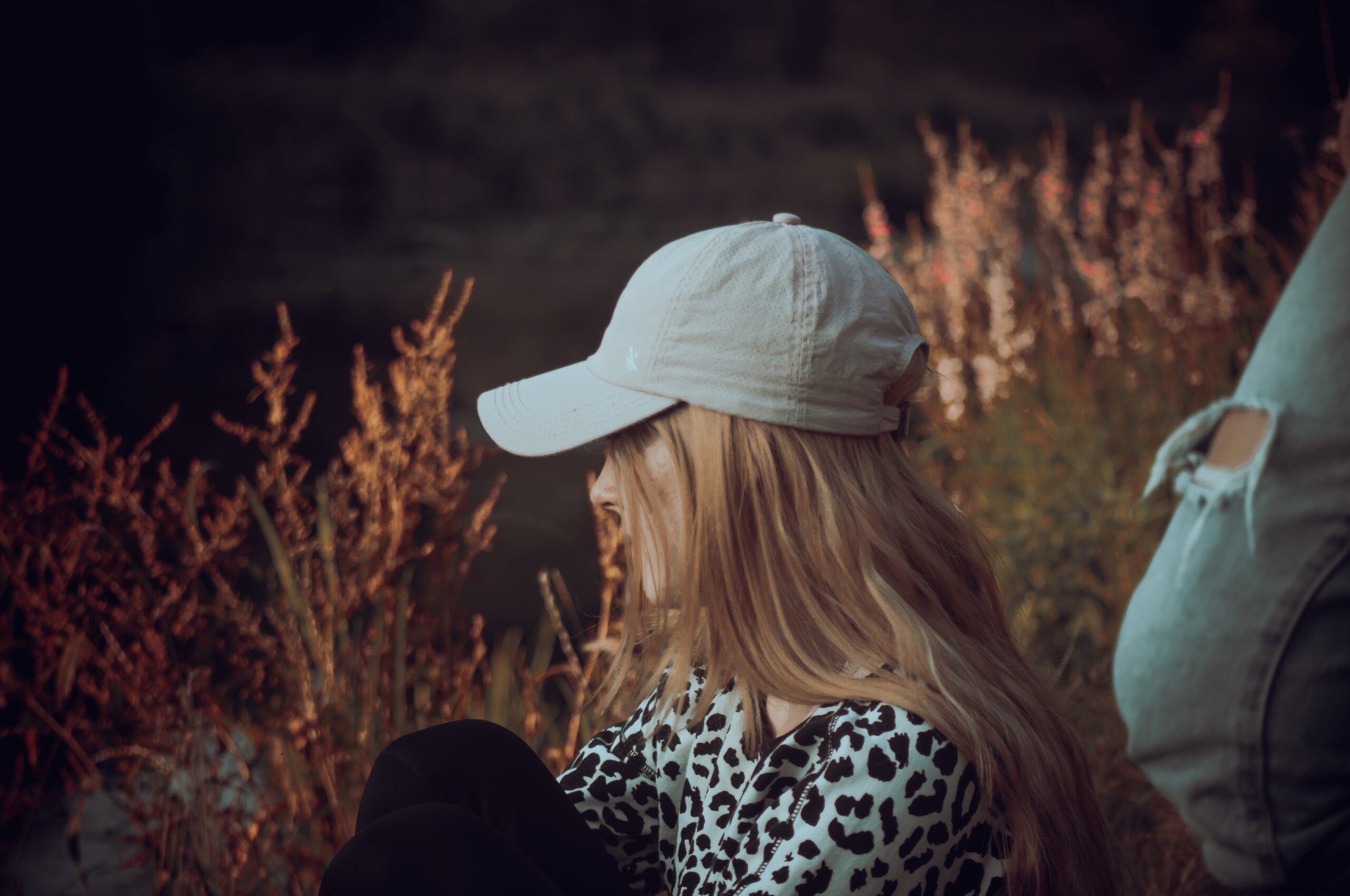 Młoda kobieta w bluzce w panterkę i czapką z daszkiem na głowie siedząca na trawie